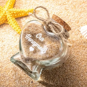 May include: A heart-shaped glass bottle filled with sand, featuring the white text Our Honeymoon and a small heart icon. The bottle is sealed with a cork stopper tied with a natural jute twine bow, positioned near a yellow starfish.