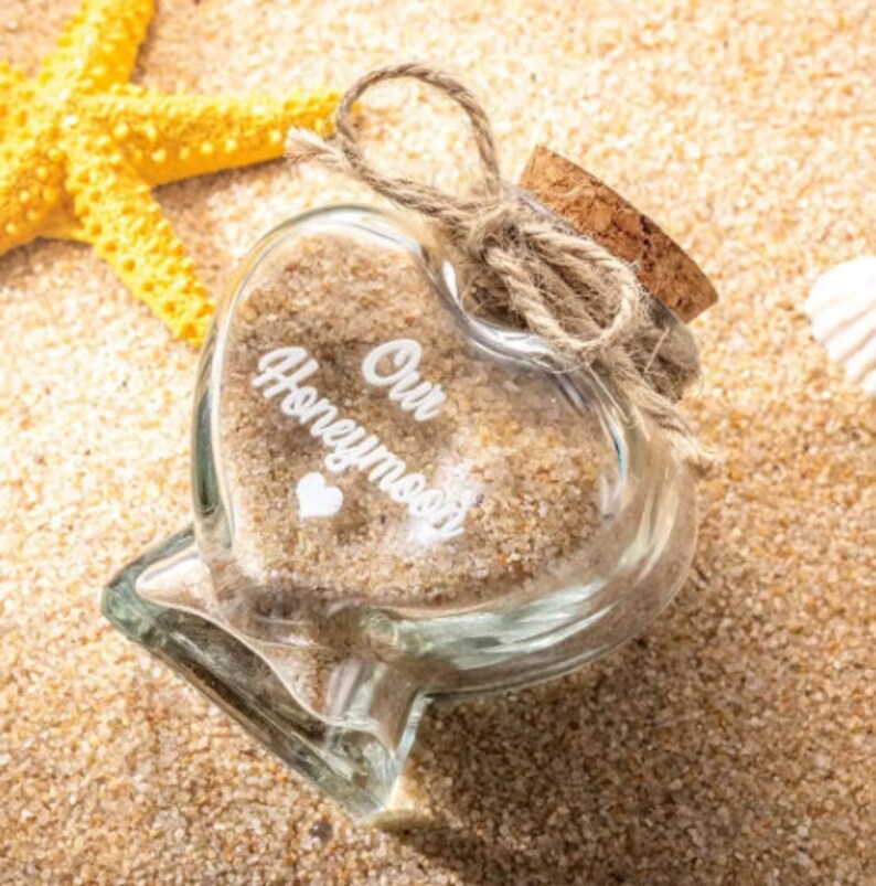 May include: A heart-shaped glass bottle filled with sand, featuring the white text Our Honeymoon and a small heart icon. The bottle is sealed with a cork stopper tied with a natural jute twine bow, positioned near a yellow starfish.