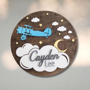 May include: A round wooden sign with a blue airplane, white clouds, yellow stars, and a yellow crescent moon. The name "Cayden Lee" is displayed in gray and white lettering. The sign is a decorative item.