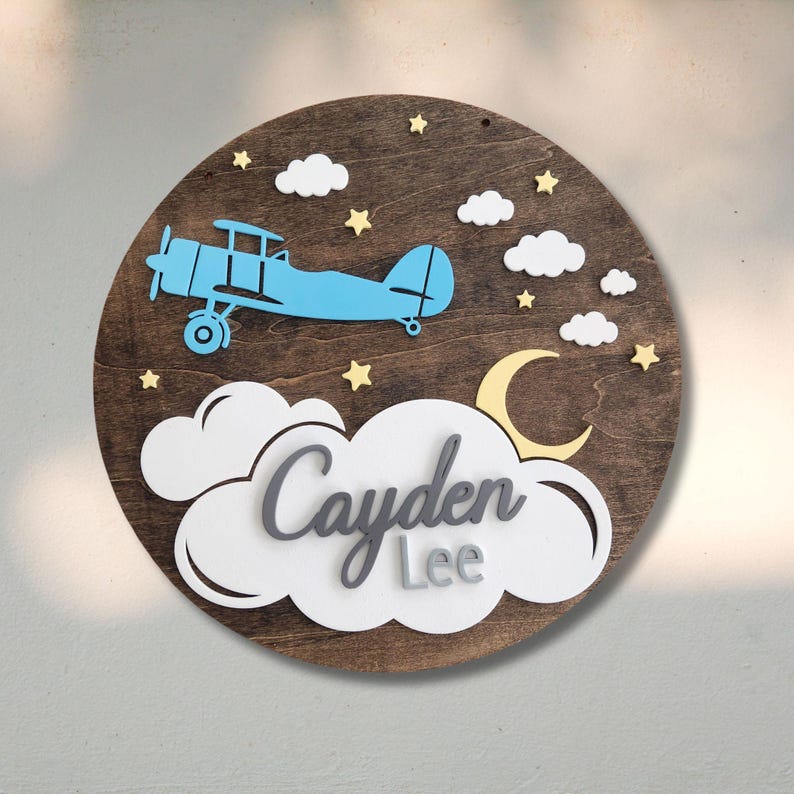 May include: A round wooden sign with a blue airplane, white clouds, yellow stars, and a yellow crescent moon. The name "Cayden Lee" is displayed in gray and white lettering. The sign is a decorative item.