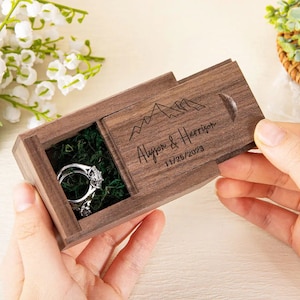 May include: A wooden ring box with a sliding lid. The lid features a mountain range design and the text "Alyson & Harrison 11/25/2023". The box is open and a silver ring is inside.
