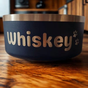 May include: A dark blue pet bowl with a metallic silver rim. The word Whiskey is printed in a light-coloured, rounded sans-serif font on the side, accompanied by two small paw print icons positioned to the right of the text.