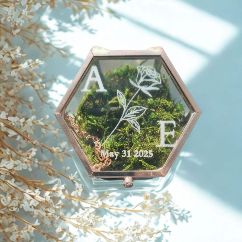K&ouml;nnte beinhalten: Eine sechseckige Glas-Ringbox mit einem ros&eacute;goldenen Metallrahmen, gef&uuml;llt mit gr&uuml;nem Moos. Der Glasdeckel zeigt wei&szlig;e gravierte Schriftz&uuml;ge, die Buchstaben A und E, eine Strichzeichnung einer Rose und das Datum May 31 2025, mit einer Goldkette.