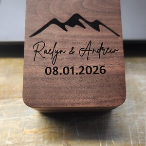 May include: A rectangular dark brown wooden box with rounded corners. The lid features a laser-engraved mountain range graphic at the top, followed by the names Raelyn & Andrew in a script font, and the date 08.01.2026 in a simple sans-serif font.