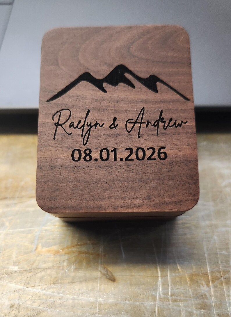 May include: A rectangular dark brown wooden box with rounded corners. The lid features a laser-engraved mountain range graphic at the top, followed by the names Raelyn & Andrew in a script font, and the date 08.01.2026 in a simple sans-serif font.
