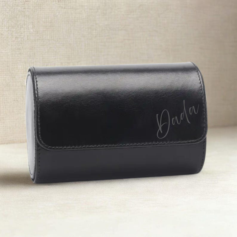 May include: A black leather watch case with a curved rectangular shape. The word "Dada" is written in cursive on the front. The case has a smooth, shiny finish and a hinged lid.