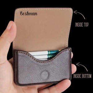 May include: A brown leather cigarette case with a tan interior, held open to reveal cigarettes. The case has the word "Bestman" printed on the inside top.