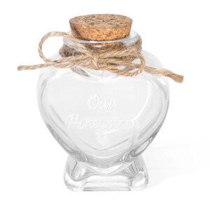 May include: A clear glass heart-shaped jar with a cork stopper and a tied jute twine bow around the neck. The front of the glass is etched with the text Our Honeymoon and a small heart symbol below the words. The jar has a flat base for stability.