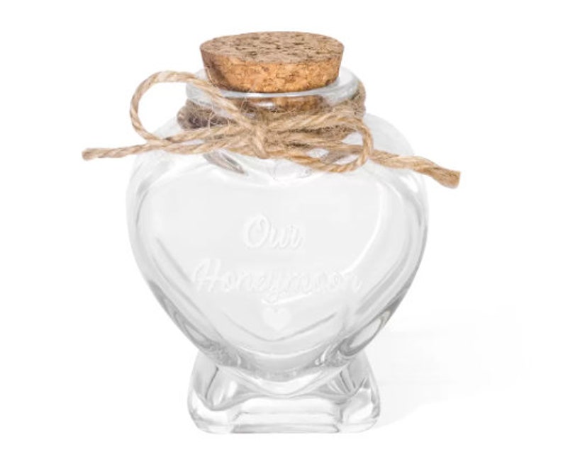 May include: A clear glass heart-shaped jar with a cork stopper and a tied jute twine bow around the neck. The front of the glass is etched with the text Our Honeymoon and a small heart symbol below the words. The jar has a flat base for stability.