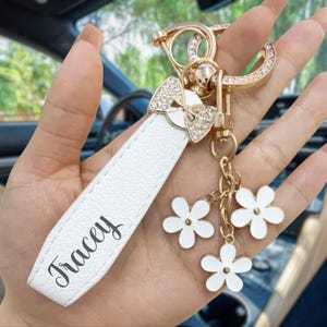Op de afbeelding: Witte leren sleutelhanger met de naam "Tracey" in sierlijke letters. De sleutelhanger heeft een goudkleurige strik met strass, een gouden sluiting en drie witte bloemvormige bedels. De achtergrond is wazig.