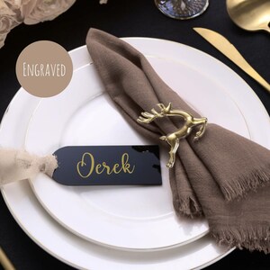 May include: A table setting featuring a gold deer napkin ring, a brown napkin, and a black name tag with "Derek" in gold. The white plate has a gold rim. A tan circle with the word "Engraved" is also visible.