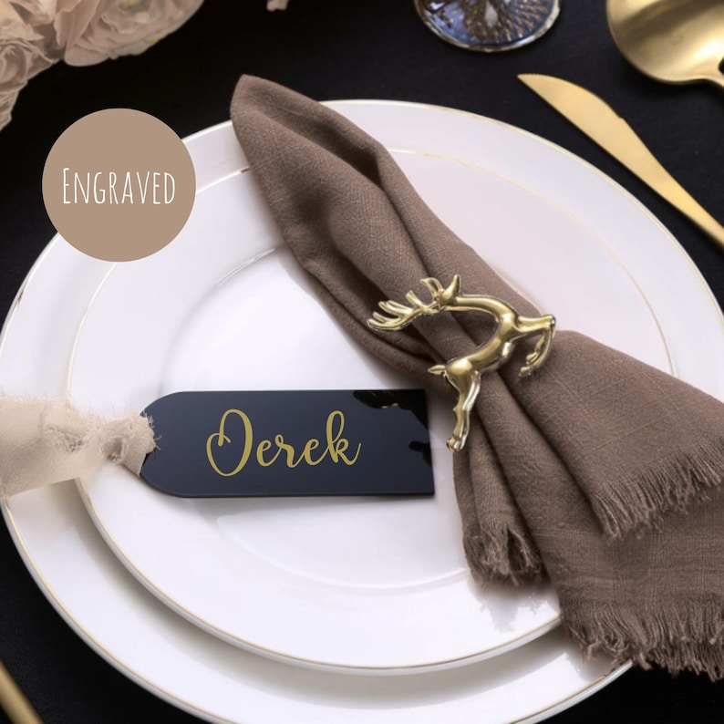 May include: A table setting featuring a gold deer napkin ring, a brown napkin, and a black name tag with "Derek" in gold. The white plate has a gold rim. A tan circle with the word "Engraved" is also visible.