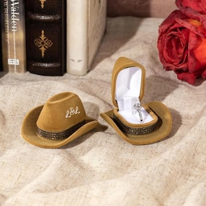 May include: Two tan velvet cowboy hat ring boxes. One box has the initials "L&L" embroidered on the front. The other box has a silver ring with a clear stone inside.
