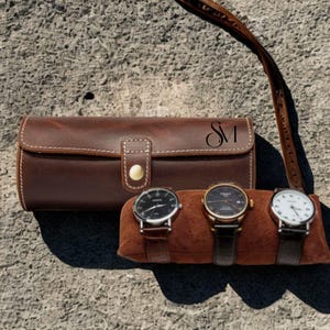May include: A brown leather watch case with white stitching and a snap closure, next to three wristwatches on a brown suede pillow. The watches have black and white faces with brown leather straps. The case has the initials "SM".