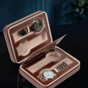 Puede incluir: Una caja de reloj de cuero marr&oacute;n con dos compartimentos, cada uno con un forro beige suave. La caja est&aacute; abierta, revelando dos relojes con correas plateadas y negras.