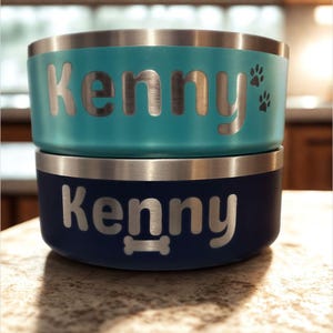 May include: Two stacked pet bowls with a metallic rim. The top bowl is turquoise with the name Kenny and two paw prints engraved. The bottom bowl is dark blue with the name Kenny and a bone shape engraved. Both bowls feature silver lettering.