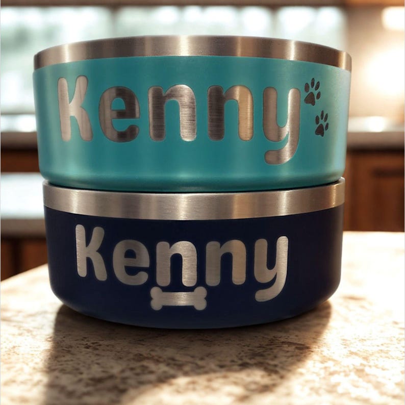 May include: Two stacked pet bowls with a metallic rim. The top bowl is turquoise with the name Kenny and two paw prints engraved. The bottom bowl is dark blue with the name Kenny and a bone shape engraved. Both bowls feature silver lettering.