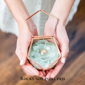 K&ouml;nnte beinhalten: Eine sechseckige Glas-Ringbox mit einem ros&eacute;goldenen Metallrahmen. Der Deckel ist mit dem Text DANIEL AND MICHELLE graviert. Das Innere ist mit hellgr&uuml;nen dekorativen Glaskieseln gef&uuml;llt und enth&auml;lt zwei silberfarbene Metallringe.