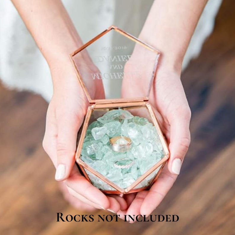 K&ouml;nnte beinhalten: Eine sechseckige Glas-Ringbox mit einem ros&eacute;goldenen Metallrahmen. Der Deckel ist mit dem Text DANIEL AND MICHELLE graviert. Das Innere ist mit hellgr&uuml;nen dekorativen Glaskieseln gef&uuml;llt und enth&auml;lt zwei silberfarbene Metallringe.