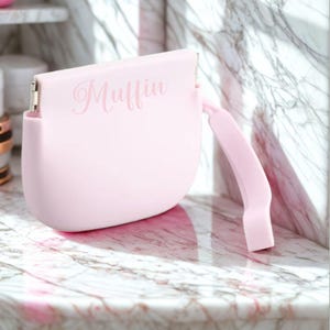 K&ouml;nnte beinhalten: Eine hellrosa Handtasche mit geschwungener Form und Griff. Das Wort "Muffin" ist in einer Schreibschrift auf der Vorderseite der Tasche geschrieben. Die Tasche steht auf einer Marmoroberfl&auml;che.