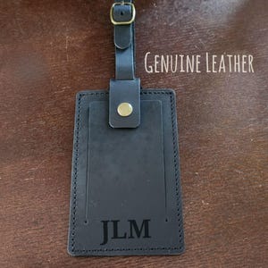 K&ouml;nnte beinhalten: Schwarzes Leder-Gep&auml;ckanh&auml;nger mit den Initialen "JLM" unten eingepr&auml;gt. Der Anh&auml;nger hat einen Riemen mit Schnalle und einen goldfarbenen Knopf. Die Worte "GENUINE LEATHER" sind oben rechts aufgedruckt.