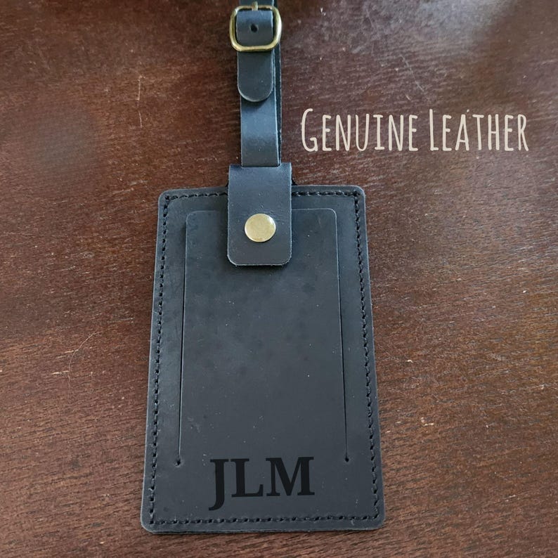 K&ouml;nnte beinhalten: Schwarzes Leder-Gep&auml;ckanh&auml;nger mit den Initialen "JLM" unten eingepr&auml;gt. Der Anh&auml;nger hat einen Riemen mit Schnalle und einen goldfarbenen Knopf. Die Worte "GENUINE LEATHER" sind oben rechts aufgedruckt.