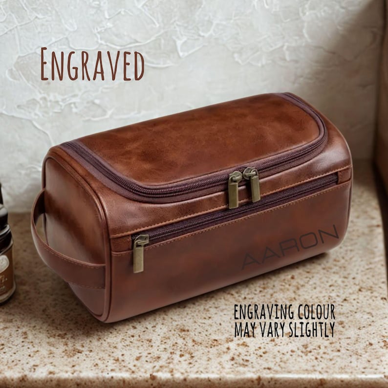 May include: A brown leather wash bag with a zipper closure and handle. The bag is engraved with the name "AARON". The text "ENGRAVED" is above the bag, and "ENGRAVING COLOUR MAY VARY SLIGHTLY" is below.