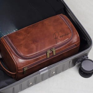 May include: A brown leather wash bag with the name "Roman" in script. The bag has a rectangular shape with a zip closure and a handle. It is inside a grey suitcase.