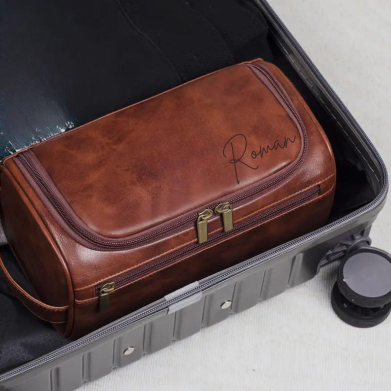 May include: A brown leather wash bag with the name "Roman" in script. The bag has a rectangular shape with a zip closure and a handle. It is inside a grey suitcase.