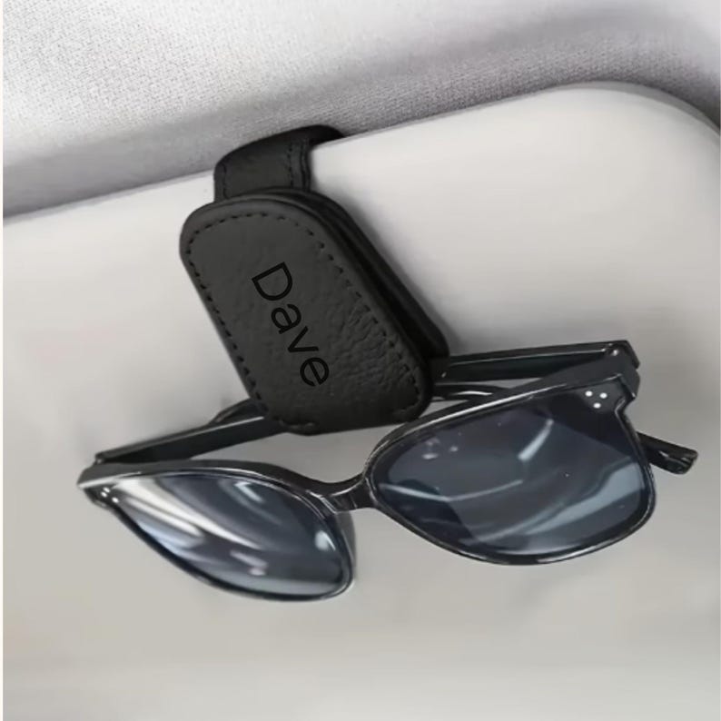 K&ouml;nnte beinhalten: Ein schwarzer Sonnenbrillenhalter aus Leder f&uuml;r die Sonnenblende im Auto mit magnetischem Clip-Mechanismus und dem Namen Dave in schwarzer Schrift auf der Vorderseite, der eine Brille mit schwarzem Rahmen und dunklen Gl&auml;sern h&auml;lt.