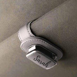 K&ouml;nnte beinhalten: Ein grauer Sonnenbrillenhalter aus Kunstleder f&uuml;r die Sonnenblende im Auto mit Magnetverschluss und einer rechteckigen Frontplatte, auf der der Name Sarah in schwarzer Schreibschrift eingraviert ist. Befestigt an einer strukturierten grauen Sonnenblende.