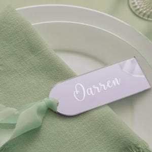 May include: A light purple name tag with the name "Darren" in white script sits on a green napkin. The napkin is on a white plate. A green ribbon is tied around the napkin. The image is a close-up of a place setting.