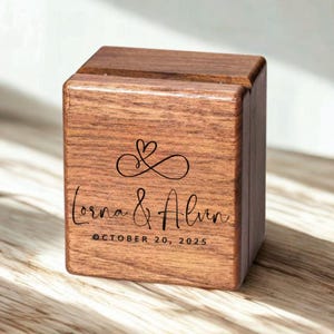 May include: A square wooden ring box with a dark brown wood grain finish. The front features a black engraved infinity symbol with a heart, the names Lorna & Alvin in script, and the date OCTOBER 20, 2025, in a clean sans-serif font.