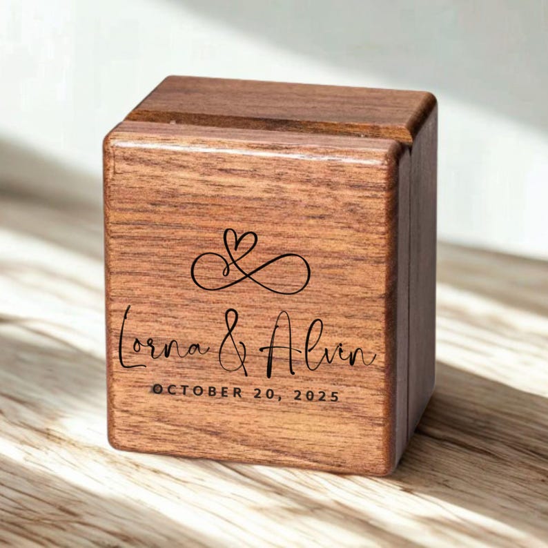 May include: A square wooden ring box with a dark brown wood grain finish. The front features a black engraved infinity symbol with a heart, the names Lorna & Alvin in script, and the date OCTOBER 20, 2025, in a clean sans-serif font.