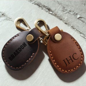 Pu&ograve; includere: Due portachiavi in pelle con hardware in ottone. Uno &egrave; marrone scuro con il nome "BRAEDON" inciso, e l'altro &egrave; marrone chiaro con le iniziali "JHC". Entrambi hanno un bordo cucito.