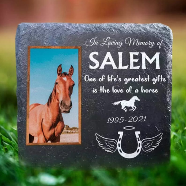 Pu&ograve; includere: Un memoriale in ardesia grigia con una foto di un cavallo marrone. La pietra ha il testo "In Loving Memory of SALEM One of life's greatest gifts is the love of a horse 1995-2021" e un ferro di cavallo con le ali.