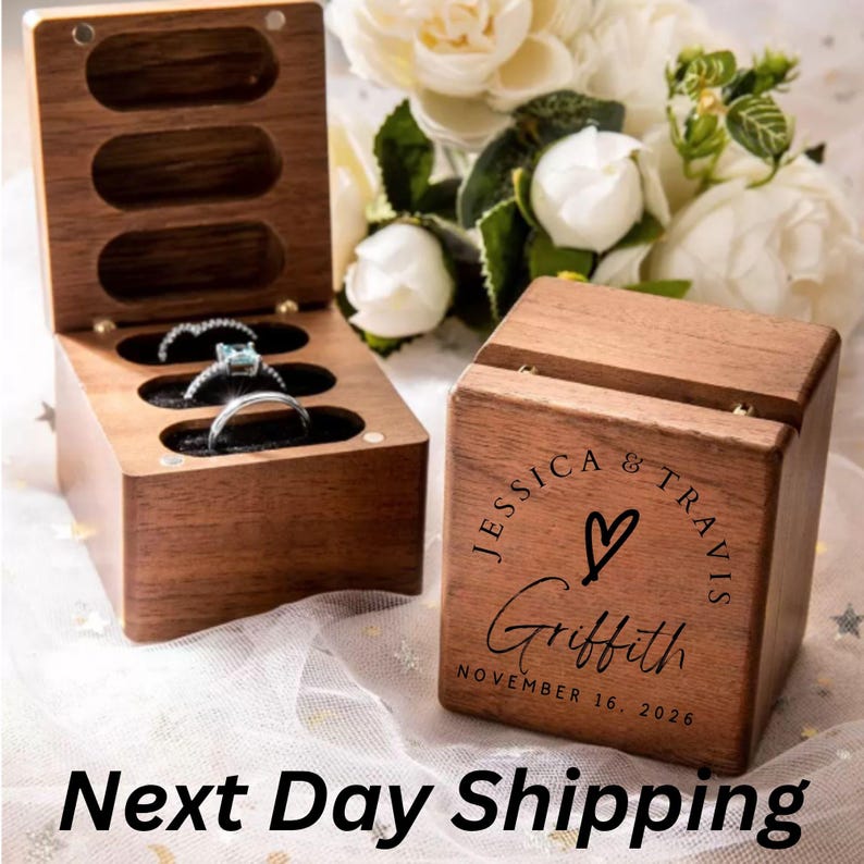 May include: A dark wood ring box with a hinged lid, featuring three oval slots containing rings. The exterior is engraved with the text JESSICA & TRAVIS, a heart symbol, the name Griffith, and the date NOVEMBER 16, 2026, in black lettering.