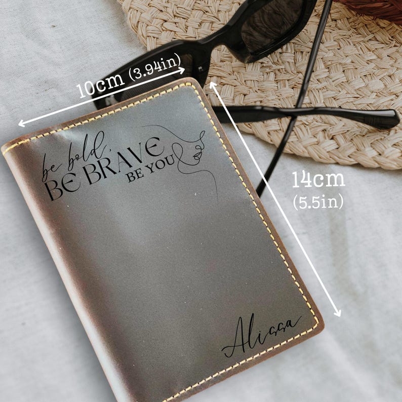 K&ouml;nnte beinhalten: Ein grauer Leder-Reisepasshalter, 14 cm x 10 cm, mit goldfarbenen N&auml;hten. Der Einband tr&auml;gt den Text "be bold BE BRAVE BE YOU" und den Namen "Ali&ccedil;&ccedil;a" in Schreibschrift sowie eine Strichzeichnung eines Gesichts.