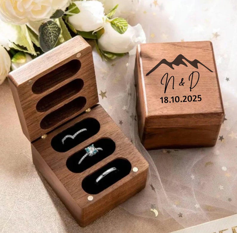 May include: A rectangular wooden ring box with a hinged lid, featuring a mountain graphic, the letters N & D, and the date 18.10.2025 engraved on the top. The interior contains three oval slots, with one ring holding a clear stone.