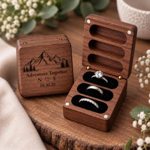 May include: A dark wood ring box with a hinged lid, featuring a mountain graphic, the text Adventure Together, initials N and E with a heart, and the date 10.30.26. The open box interior has three black velvet slots holding three silver rings.