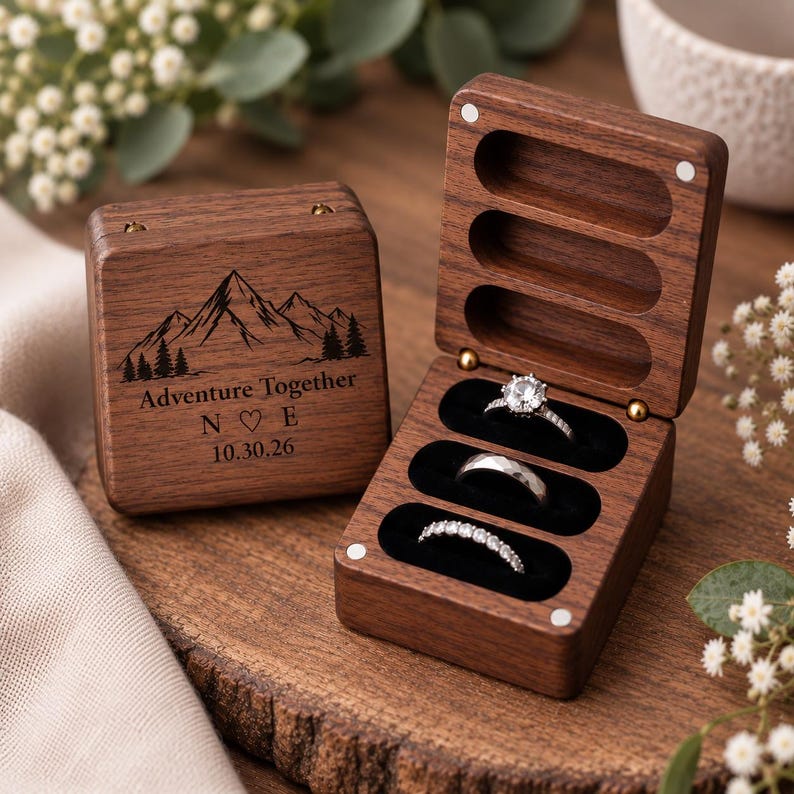 May include: A dark wood ring box with a hinged lid, featuring a mountain graphic, the text Adventure Together, initials N and E with a heart, and the date 10.30.26. The open box interior has three black velvet slots holding three silver rings.