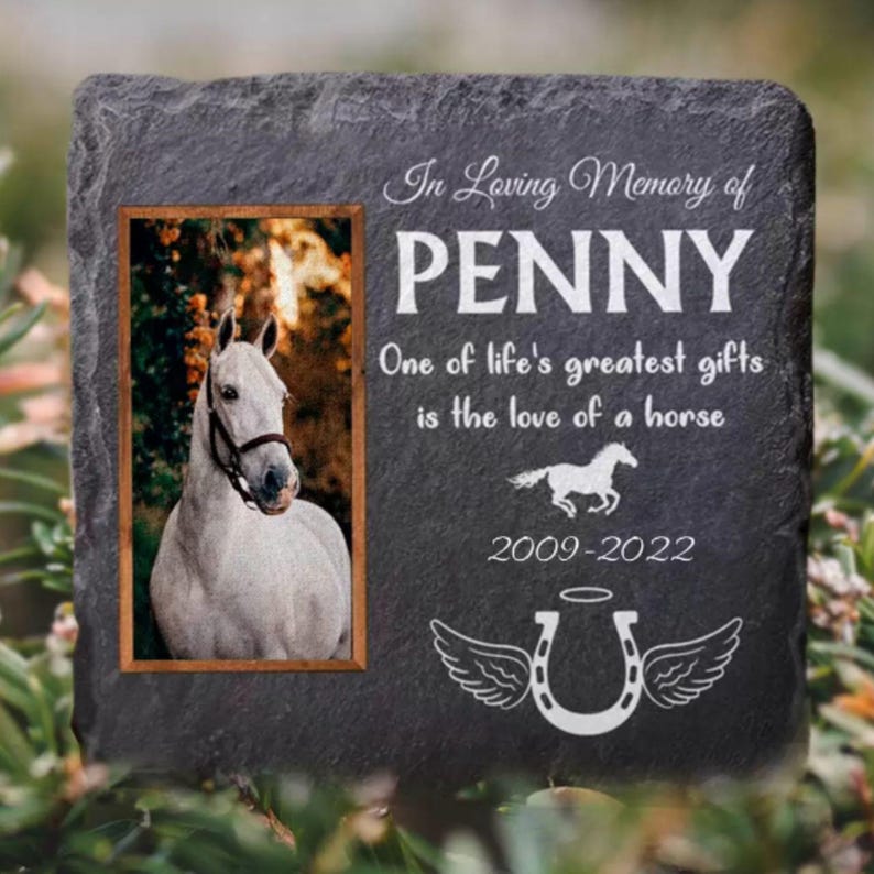 Pu&ograve; includere: Un memoriale in ardesia grigia con una foto di un cavallo bianco. La pietra ha il testo "In Loving Memory of PENNY One of life's greatest gifts is the love of a horse 2009-2022" e un ferro di cavallo con le ali.