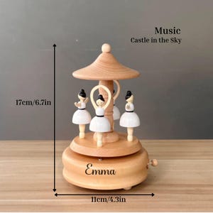 Puede incluir: Una caja de m&uacute;sica de carrusel de madera con cuatro figuras de bailarinas con vestidos blancos. La caja de m&uacute;sica mide 17 cm de alto y 11 cm de ancho, con el nombre "Emma" en la base. Tambi&eacute;n se ve el texto "Music Castle in the Sky".