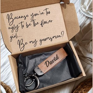 May include: A brown gift box containing a leather keychain engraved with the name "David". The box lid has the text: "Because you're too ugly to be the flower girl... Be my groomsmen?"