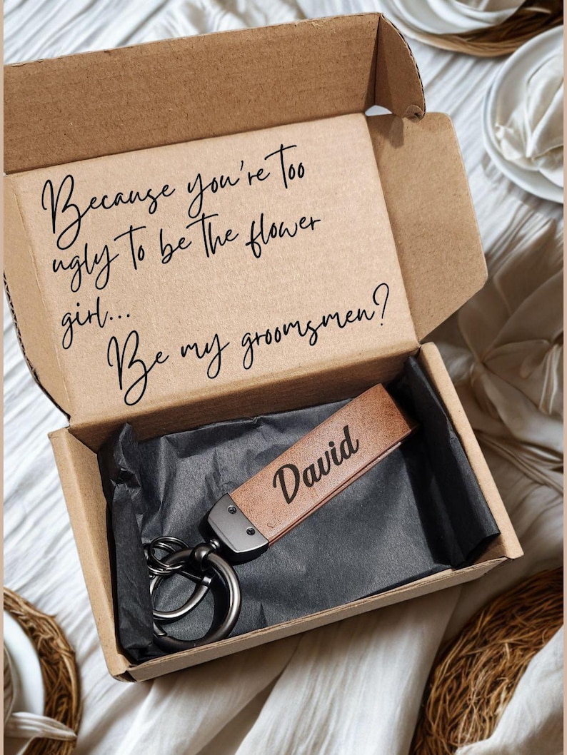 May include: A brown gift box containing a leather keychain engraved with the name "David". The box lid has the text: "Because you're too ugly to be the flower girl... Be my groomsmen?"