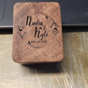May include: A rectangular wooden box with rounded corners featuring a laser-engraved design of a floral wreath surrounding the text Nadia & Kyle and the date JUNE 20, 2021. The wood grain is visible across the surface of the lid.