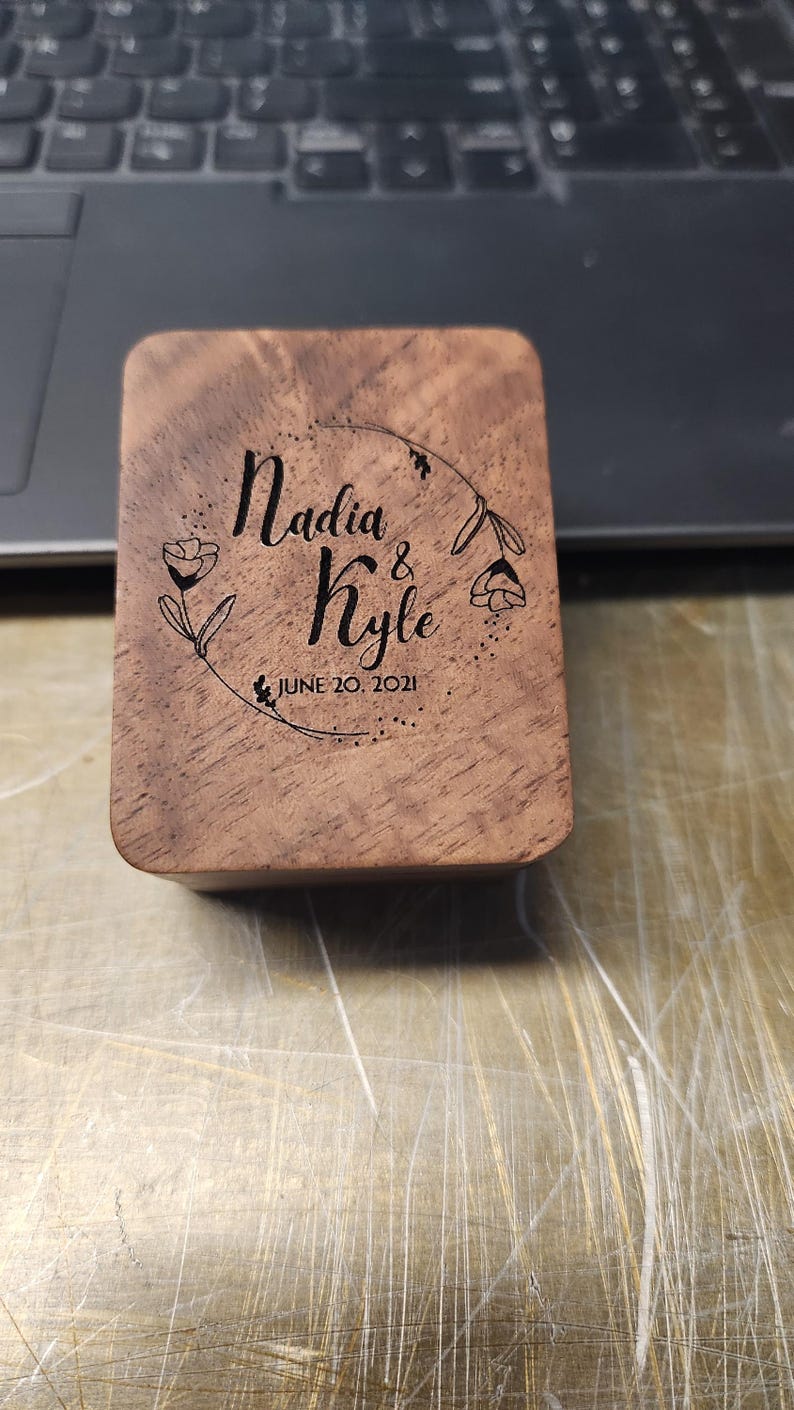 May include: A rectangular wooden box with rounded corners featuring a laser-engraved design of a floral wreath surrounding the text Nadia & Kyle and the date JUNE 20, 2021. The wood grain is visible across the surface of the lid.