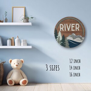 May include: A round wooden sign with the word "RIVER" in white, featuring a mountain and forest scene. Available in 12, 14, and 16 inch sizes. The sign has a rustic, natural aesthetic, suitable for home decor.