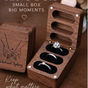 May include: A rectangular dark wood ring box with a hinged lid, featuring a laser-engraved illustration of two hands holding each other and the text 'ichel and Sydney'. The interior has three black velvet slots holding three silver rings.