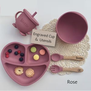 May include: A rose-colored silicone baby feeding set, including a heart-shaped divided plate, bowl, cup with lid, spoon, and fork. The spoon and cup are engraved with a name. The plate contains berries and cookies.